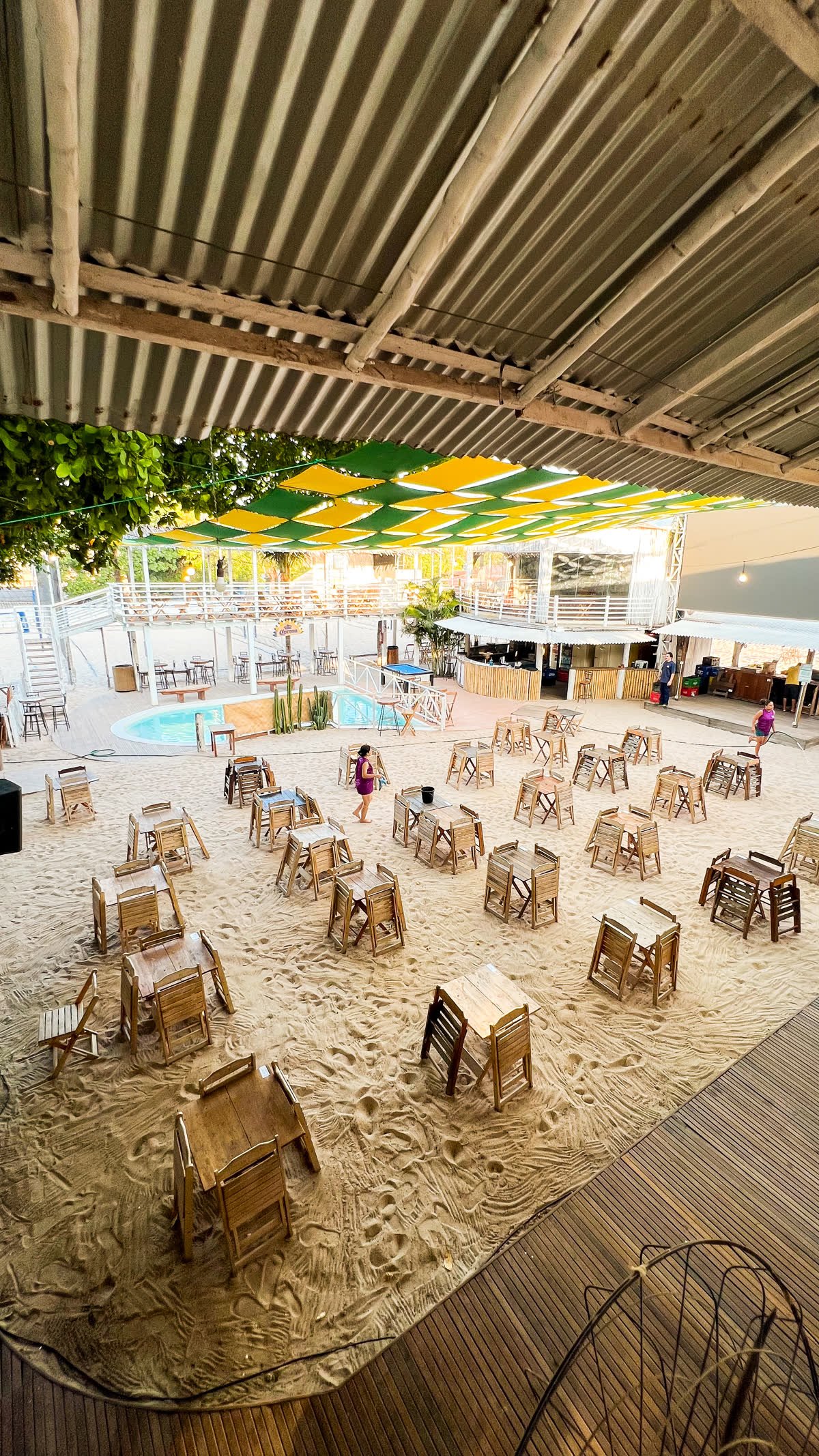Vista do alto do Rio Sand Club, com mesas e cadeiras de madeira na areia, piscina e toldo verde e amarelo ao fundo