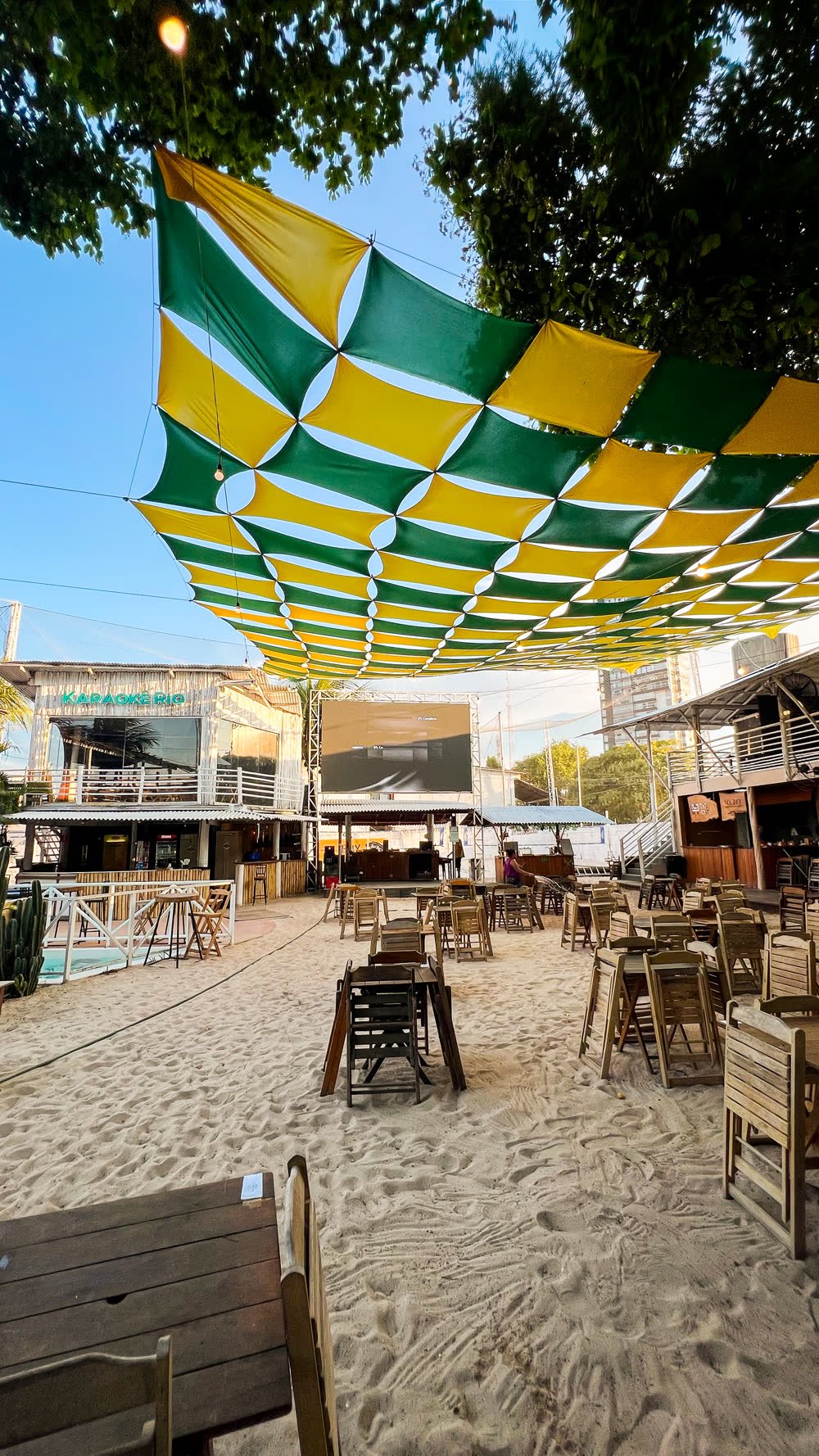 Espaço aberto do Rio Sand Club, com toldo verde e amarelo sobre as mesas na areia e o telão ao fundo