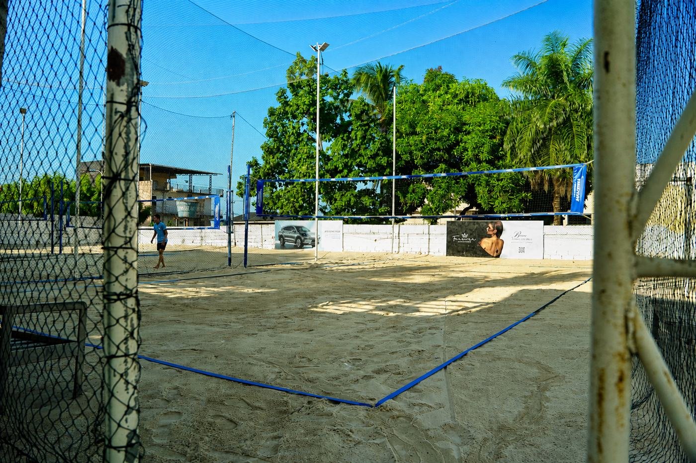 Quadra de areia do Rio Sand Club cercada por palmeiras, com jogador atravessando a areia